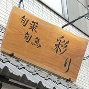 あたたかみのある木製の看板と清潔感あふれる白暖簾が迎える【旬彩旬魚　彩り】。店の顏ともいえるその文字は、須田さんが一文字一文字、丁寧に書き上げたもの。多彩な才能に驚きます。