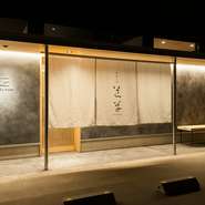 店主が友人の設計士に依頼したというスタイリッシュな建物が印象的。炭入りモルタルや温かみのある照明が、洗練された和モダンな雰囲気を演出しています。店のロゴが描かれた暖簾が目印です。