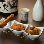 左から、ホタルイカの酢味噌がけ、ゴボウの穴子巻き、キスと大葉の梅肉のせ。和の魅力溢れる串カツのセットは、日本酒や焼酎が進むこと間違いなしです。（撮影日の一例。）