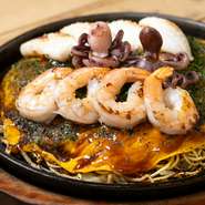 広島のスタンダードなお好み焼「肉玉そば」に、イカ・タコ・エビを乗せたデラックス。生麺のもっちりした食感も絶妙で、野菜の水分で蒸すように焼き上げます。トッピングの追加もオススメ。
※そばをうどんに変更可